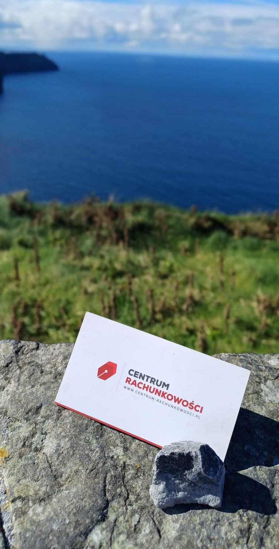 Wizytówka z logo 'Centrum Rachunkowości' na tle kamiennego murku z widokiem na morze i zielone wzgórza w oddali.