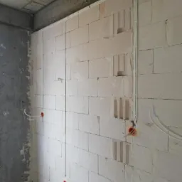 Wewnętrzna ściana z betonu komórkowego w trakcie układania przewodów elektrycznych, widoczne puszki instalacyjne i peszle prowadzone po powierzchni ściany.