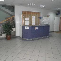 Wnętrze placówki medycznej: recepcja z niebieskim blatem, informacjami za szybą, schody w tle i donica z rośliną obok.