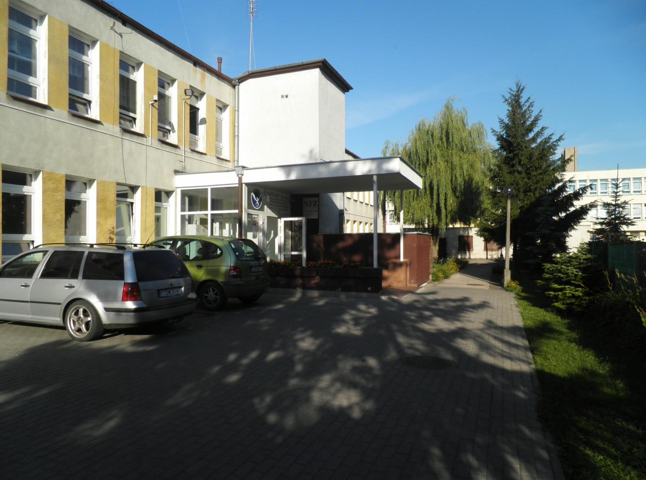 Budynek placówki medycznej z parkingiem, widoczny znak NZOZ nad wejściem.