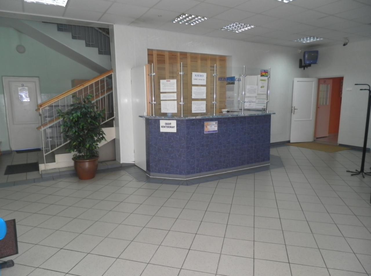 Wnętrze placówki medycznej: recepcja z niebieskim blatem, informacjami za szybą, schody w tle i donica z rośliną obok.