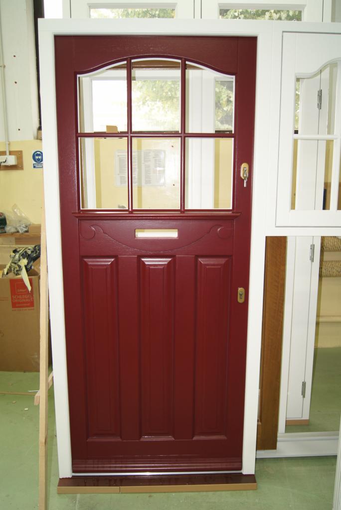 Ciemnoczerwone drzwi zewnętrzne z szybami w górnej części i skrzynką na listy, widoczne w warsztacie obok białych drzwi.
