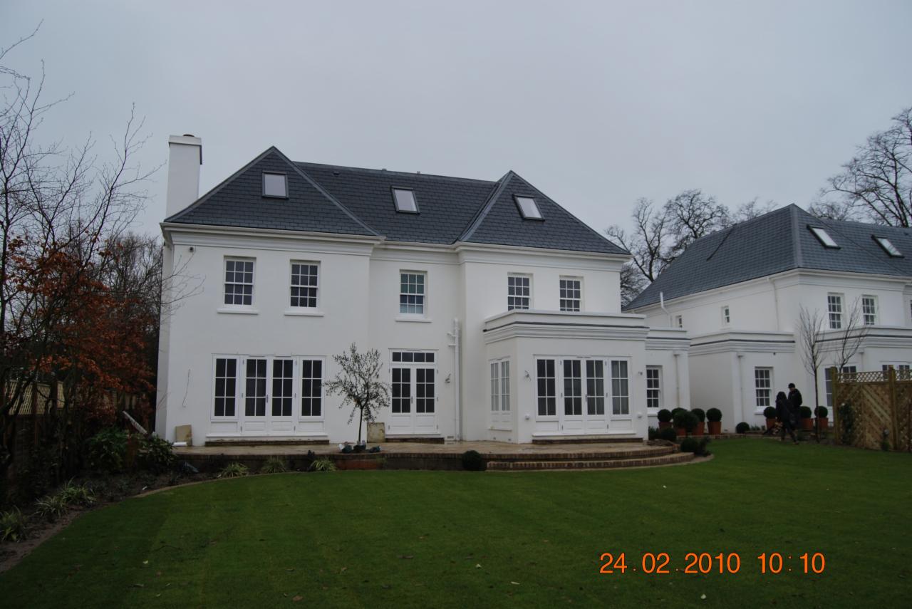 Elewacja dwukondygnacyjnego, białego domu z szarym dachem i licznymi oknami oraz przeszklonymi drzwiami tarasowymi, widok z zadbanego trawnika w pochmurny dzień.