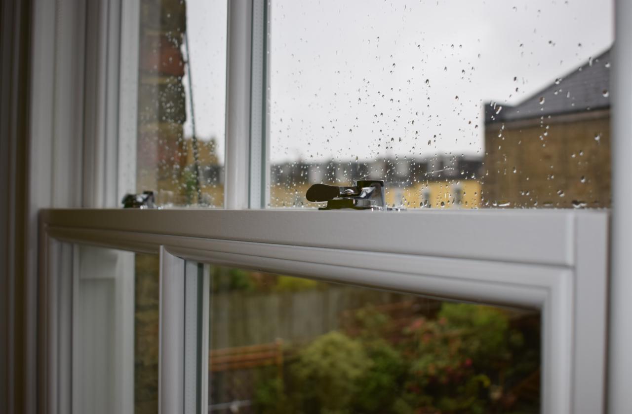Zamknięte białe okno z kroplami deszczu na szybie, widok na budynek i zieleń na zewnątrz, metalowy zatrzask okienny w pozycji zamkniętej.