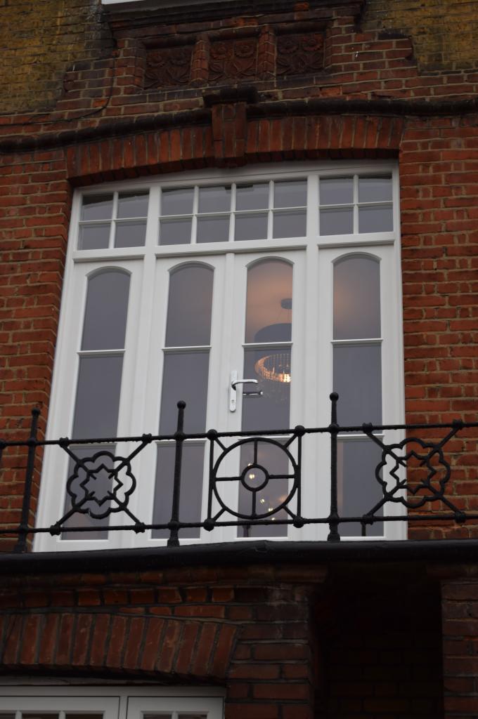 Białe okno balkonowe z czarną, ozdobną balustradą na tle ceglanej fasady budynku, wewnątrz widoczny żyrandol.