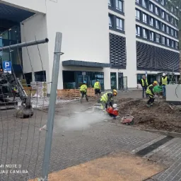 Pracownicy budowlani w żółtych kamizelkach tną kostkę brukową na placu budowy przed nowoczesnym budynkiem biurowym, z widocznym mini-ekskawatorem i materiałami budowlanymi w tle.