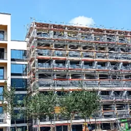 Wielopiętrowy budynek w trakcie budowy z rusztowaniami, widoczne elementy konstrukcyjne i robotnicy w oddali, obok gotowy budynek z balkonami i szklanymi balustradami, zielone drzewa na pierwszym...