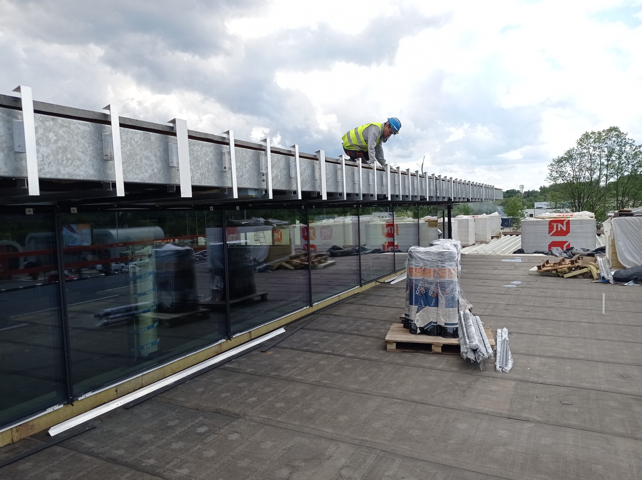 Pracownik w kasku i kamizelce odblaskowej pracuje na dachu budynku z elementami metalowej konstrukcji, w tle zapakowane materiały budowlane i odbicie otoczenia w szklanej fasadzie.
