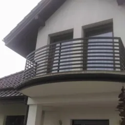 Nowoczesna, czarna balustrada balkonowa z poziomych prętów, zamontowana na balkonie domu z ciemną dachówką i ozdobnym elementem wiszącym.