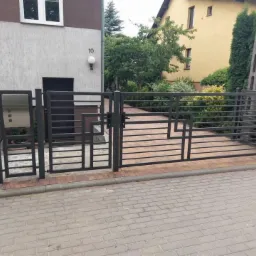 Nowoczesna, antracytowa brama wjazdowa z poziomymi prętami i geometrycznymi zdobieniami, zamontowana przed domem z numerem 10, widoczna z poziomu kostki brukowej.