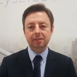 Portret mężczyzny w garniturze i krawacie na tle rozmytego krajobrazu miejskiego w odcieniach szarości.