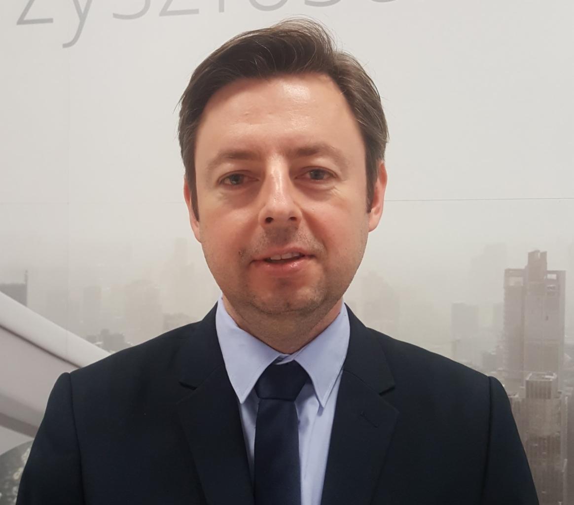 Portret mężczyzny w garniturze i krawacie na tle rozmytego krajobrazu miejskiego w odcieniach szarości.