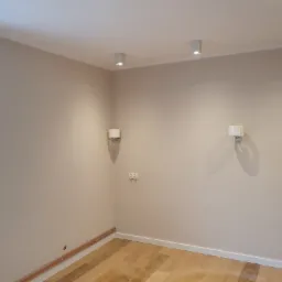 Pomalowane na beżowo wnętrze pokoju z drewnianą podłogą, białymi listwami przypodłogowymi, grzejnikiem pod oknem, miedzianymi rurami przy ścianie, gniazdkami elektrycznymi i kinkietami.