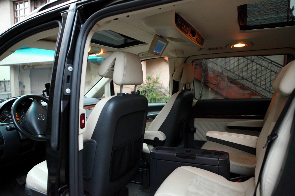 Wnętrze luksusowego, czarnego vana Mercedes z beżowymi skórzanymi siedzeniami, widok z otwartych drzwi kierowcy, podsufitka z monitorem i oknem dachowym.