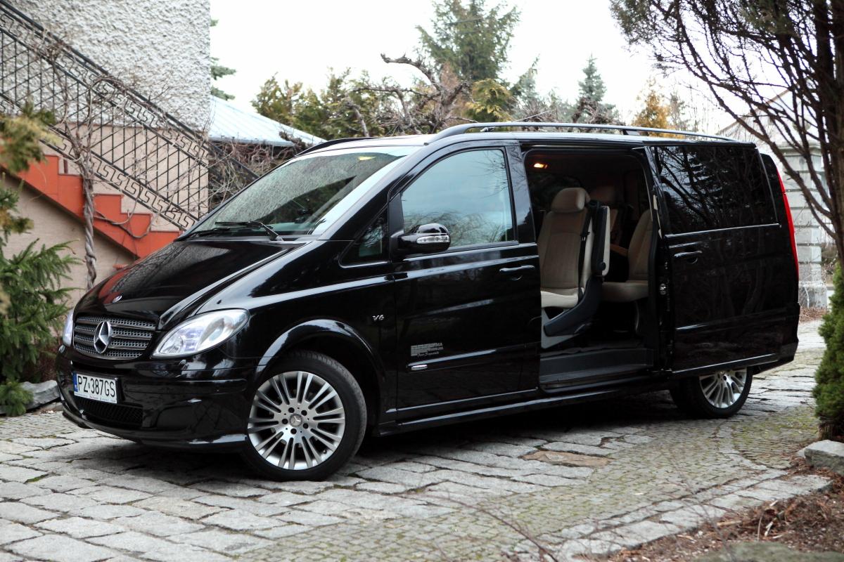 Czarny Mercedes Viano z otwartymi drzwiami przesuwnymi, zaparkowany na brukowanej drodze przed budynkiem z jasną elewacją i ozdobnymi schodami.