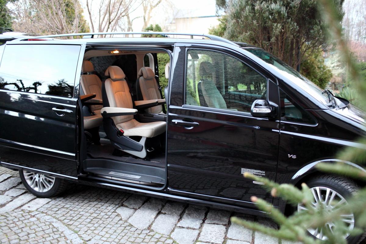 Czarny van z otwartymi drzwiami, ukazujący jasne skórzane fotele z podłokietnikami, zaparkowany na brukowanej drodze, z widocznym logo 'V6' na błotniku.