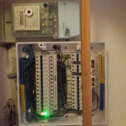 Otwarta skrzynka elektryczna z widocznymi bezpiecznikami, przewodami i listwami zaciskowymi, zamontowana obok licznika energii elektrycznej RWE na ścianie wewnątrz budynku.