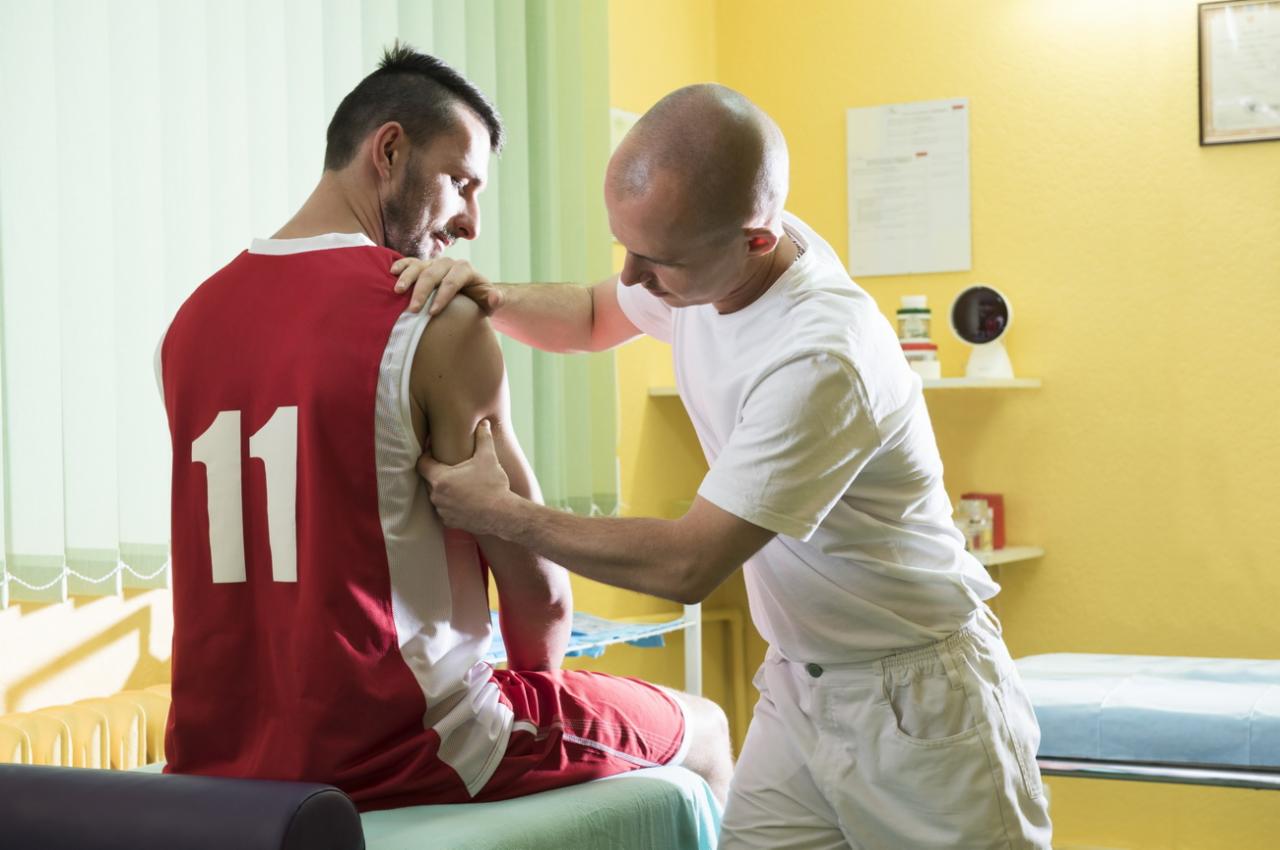 Mężczyzna w koszulce sportowej numer 11 poddawany terapii manualnej ramienia przez terapeutę w białym uniformie, w gabinecie z żółtymi ścianami.