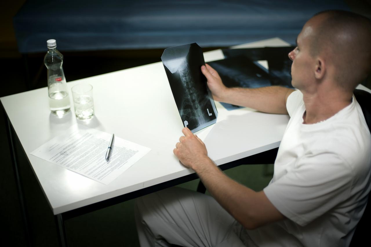 Mężczyzna w białym ubraniu siedzi przy biurku i analizuje zdjęcie rentgenowskie kręgosłupa, obok leżą inne zdjęcia, woda i dokumenty.