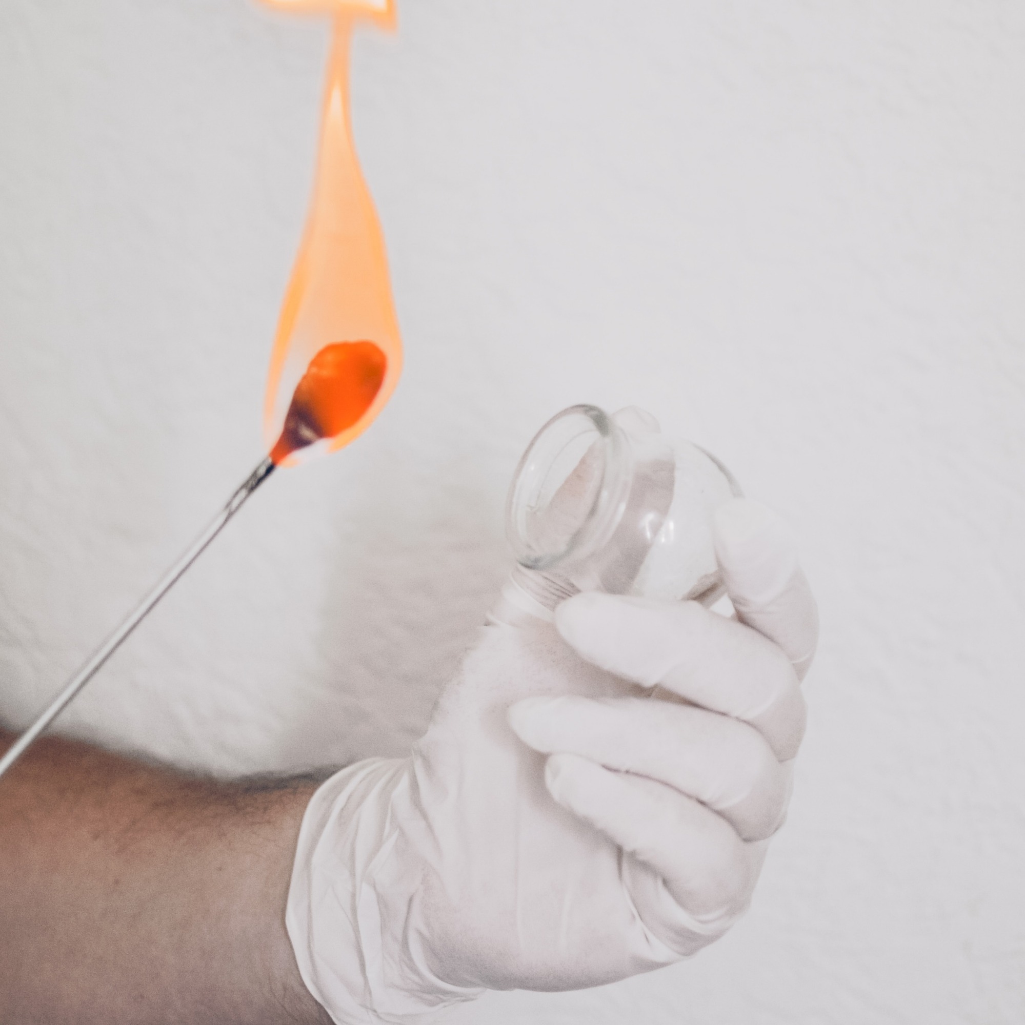 Szklana bańka do masażu ogniowego ogrzewana płomieniem, trzymana w dłoni w białej rękawiczce na tle białego prześcieradła.