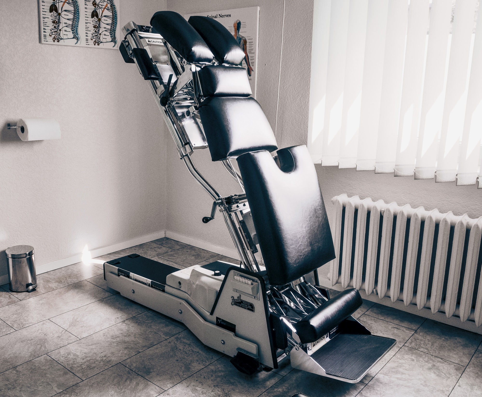 Profesjonalny stół do terapii manualnej z regulowanymi segmentami, chromowaną ramą i czarną tapicerką, ustawiony w jasnym gabinecie z grzejnikiem i plakatem anatomicznym.