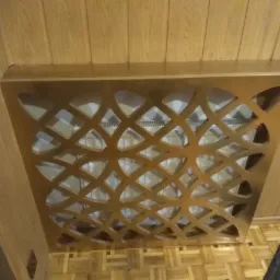 Drewniana osłona grzejnika z ażurowym wzorem na tle boazerii i parkietu.