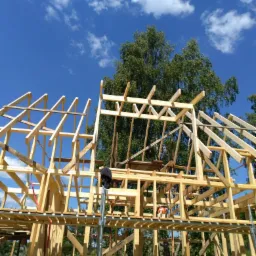 Konstrukcja szkieletowa domu w trakcie budowy, widok z dołu na drewniane elementy i rusztowanie, błękitne niebo w tle.