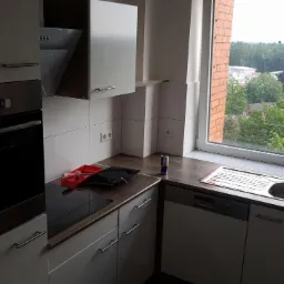 Kąt ujęcia na fragment kuchni z białymi szafkami, piekarnikiem z etykietą energetyczną, zlewem i widokiem na zieleń za oknem; na blacie leżą akcesoria malarskie i puszka napoju energetycznego.