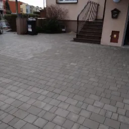 Ułożona szara kostka brukowa na podjeździe przed domem z numerem 20, widoczny kosz na śmieci i wejście do budynku ze schodami.