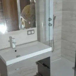 Nowoczesna, częściowo wykończona łazienka z umywalką nablatową, lustrem z oświetleniem i kabiną prysznicową typu walk-in. Widoczne niedokończone szafki pod umywalką i narzędzia na podłodze.