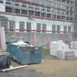 Budynek wielopiętrowy w trakcie prac remontowych, otoczony rusztowaniem i kontenerem na odpady, z widocznymi materiałami budowlanymi.