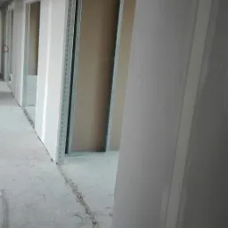 Korytarz w trakcie wykańczania, widoczne ściany z płyt kartonowo-gipsowych na metalowym stelażu, częściowo zaszpachlowane łączenia, surowy beton na podłodze, widoczne kable i drobne odpady budowlane.