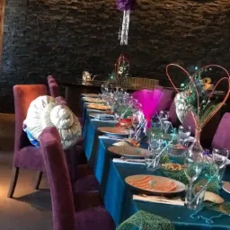 Uroczysty stół z turkusowym obrusem i fioletowymi krzesłami, dekoracje w stylu morskim, talerze z przekąskami, szklanki i sztućce, wiszące ozdoby z bibuły i frędzli, kamienna ściana w tle.