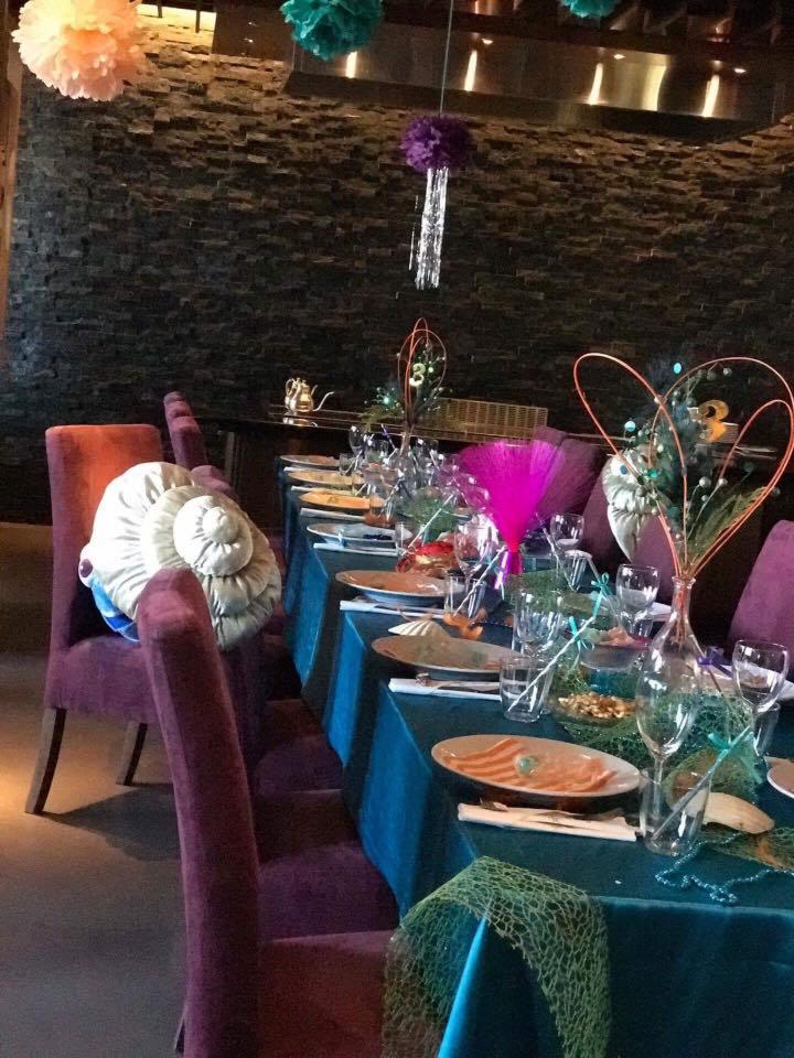 Uroczysty stół z turkusowym obrusem i fioletowymi krzesłami, dekoracje w stylu morskim, talerze z przekąskami, szklanki i sztućce, wiszące ozdoby z bibuły i frędzli, kamienna ściana w tle.