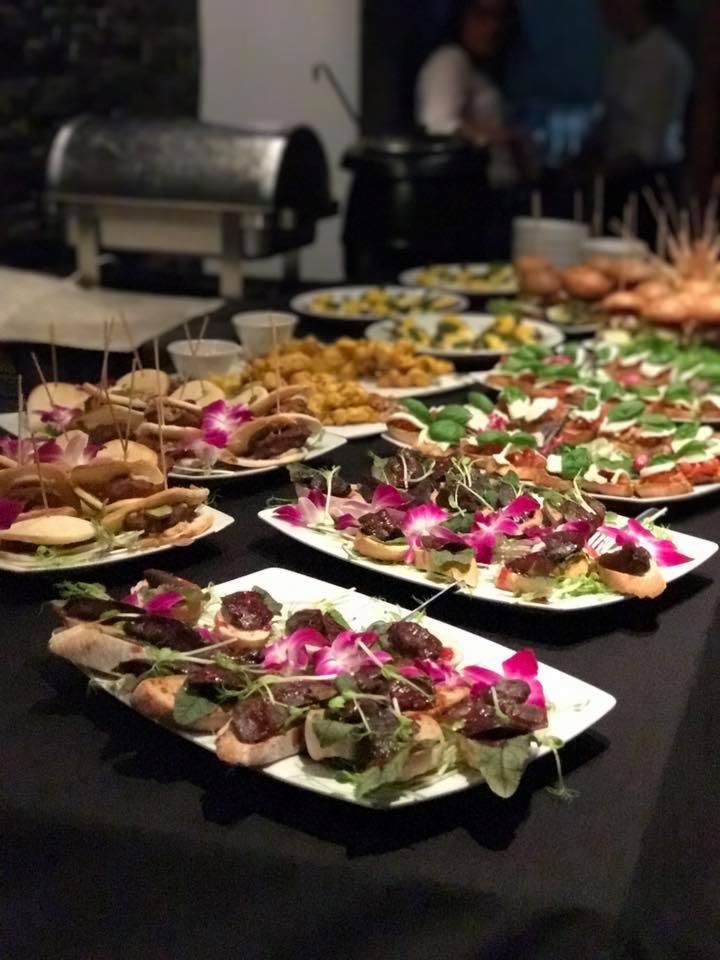 Stół z różnorodnymi przekąskami na białych talerzach, udekorowanymi jadalnymi kwiatami, w tle podgrzewacze i obsługa.