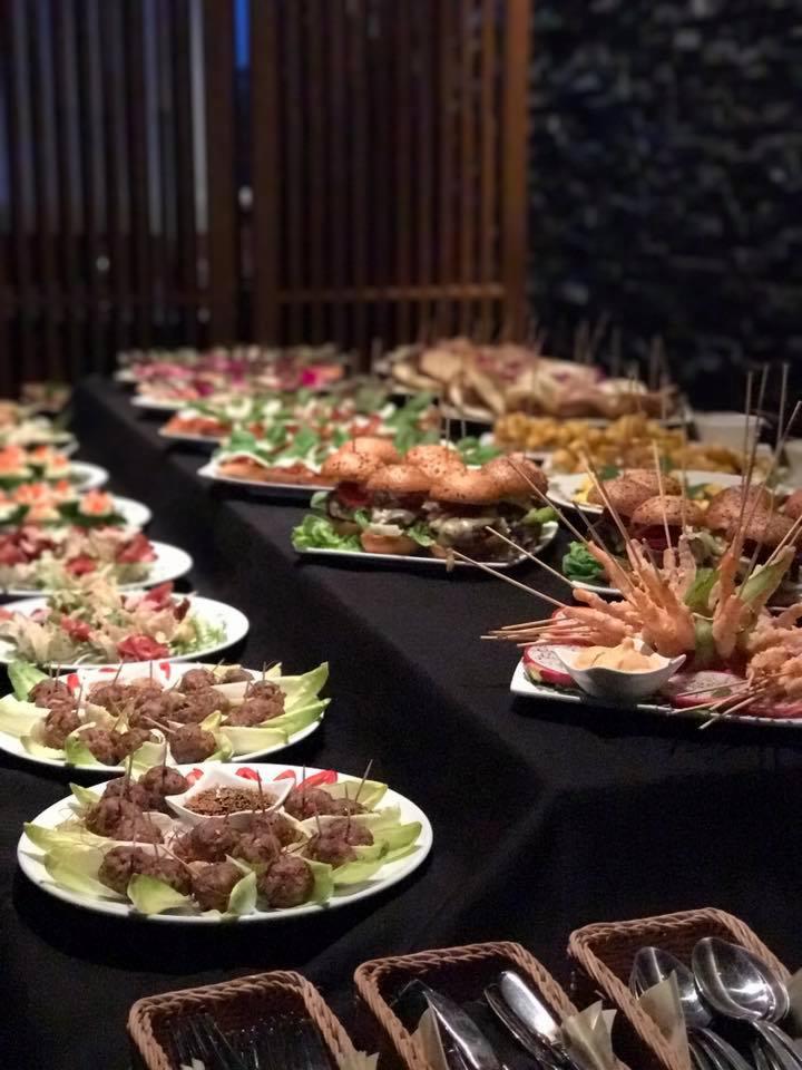 Stół z czarnym obrusem zastawiony różnorodnymi przekąskami na białych talerzach, w tym mini-burgery z sezamem, szaszłyki z krewetek i mięsne koreczki na liściach sałaty, uzupełniony sztućcami...