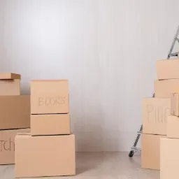 Stos kartonowych pudeł z napisem 'Kitchen', 'Books' i 'Pillow' w pokoju, z drabinką opartą o ścianę, przygotowanych do przeprowadzki.