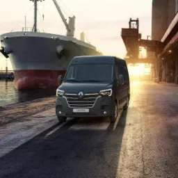 Czarny Renault Master na tle portu z dużym statkiem towarowym i dźwigami, obok stoi pomarańczowy bus.