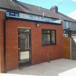 Nowo dobudowana, czerwona, ceglana przybud&oacute;wka do domu z brązowymi oknami i drzwiami, wyłożona jasną kostką brukową.