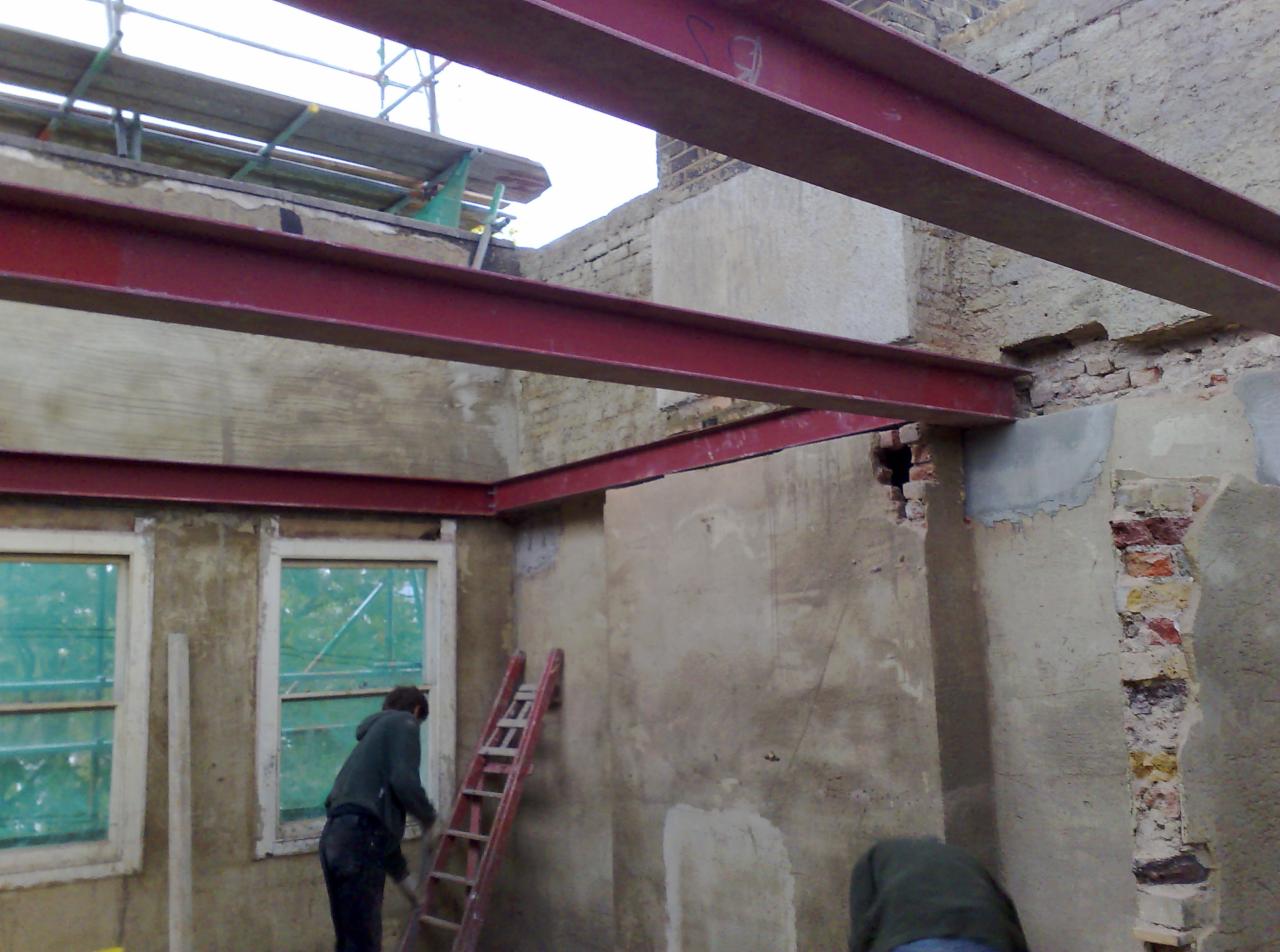 Wnętrze remontowanego budynku z widocznymi stalowymi belkami nośnymi, fragmentami otynkowanych ścian, oknami zabezpieczonymi folią i pracownikami przy pracy.