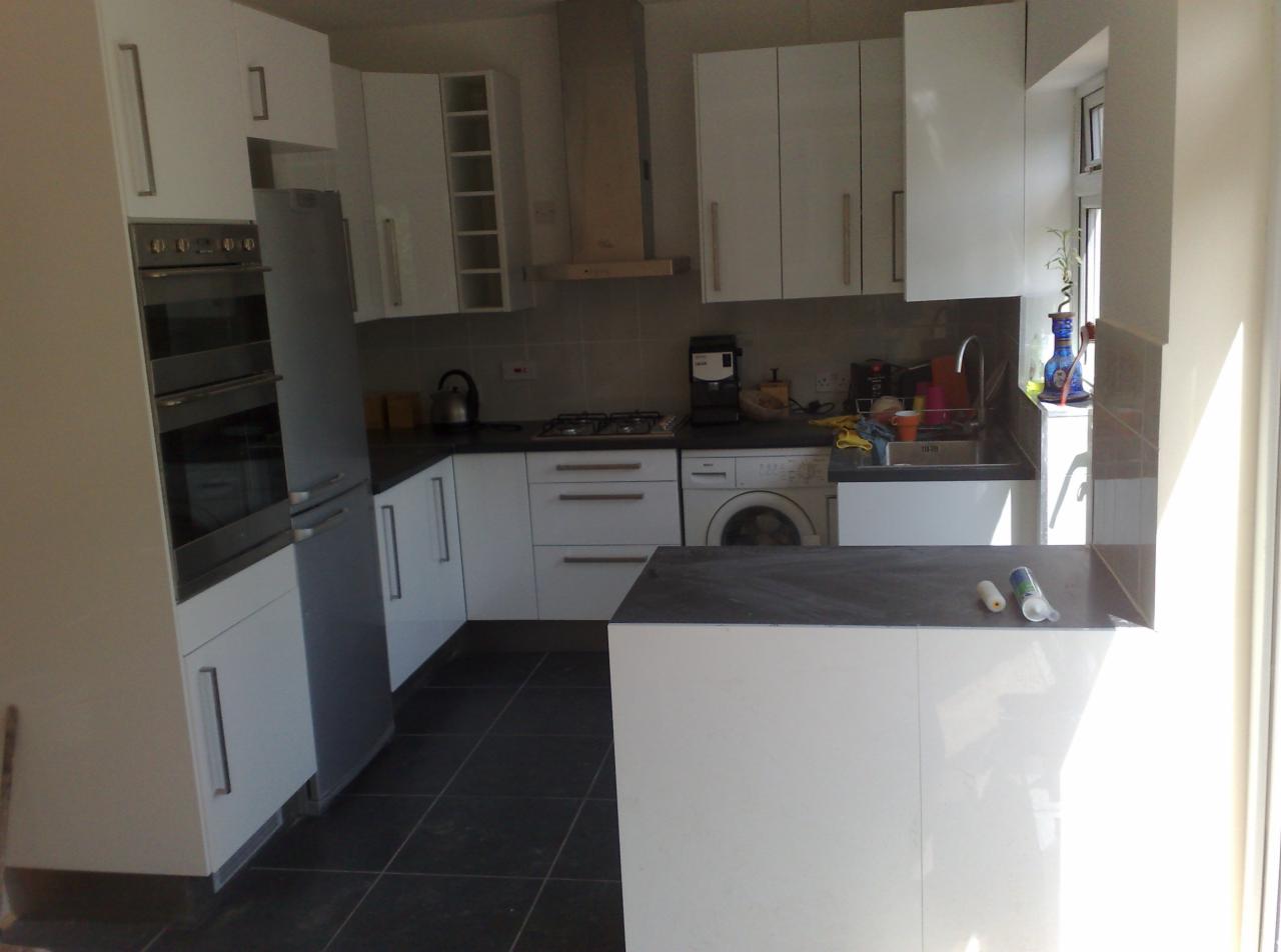 Jasna kuchnia z białymi szafkami, czarnym blatem, sprzętami AGD i ciemną podłogą z płytek; widoczna pralka w zabudowie kuchennej.