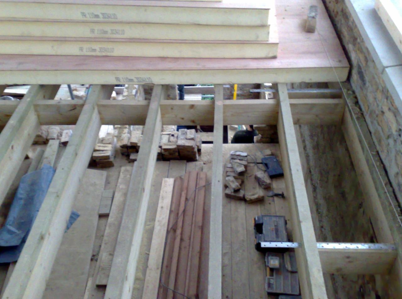 Wnętrze w trakcie budowy z drewnianą konstrukcją stropu, widoczne cegły, narzędzia i fragment ściany z kamienia.