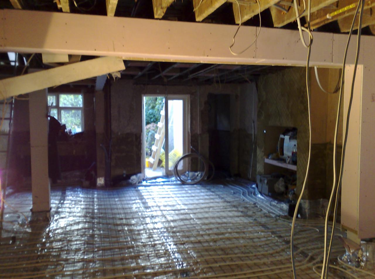 Instalacja ogrzewania podłogowego w trakcie remontu, widoczne rury grzewcze na podłodze, surowe ściany, okno i drzwi balkonowe w tle, elementy instalacji elektrycznej pod sufitem.