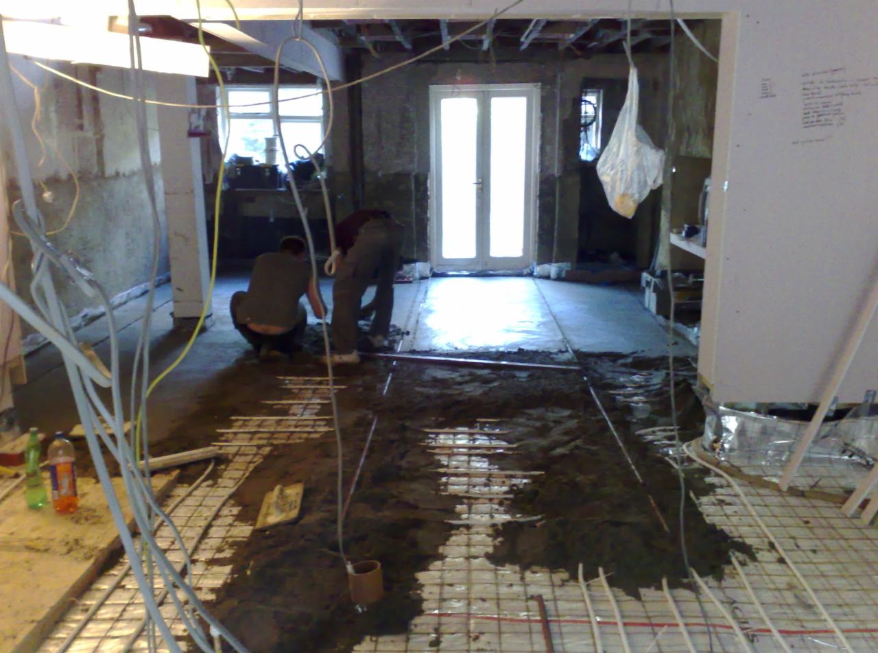Dwaj mężczyźni w trakcie wylewania posadzki z ogrzewaniem podłogowym na siatce metalowej, widoczne kable elektryczne, butelki z napojami i surowe ściany w trakcie remontu.
