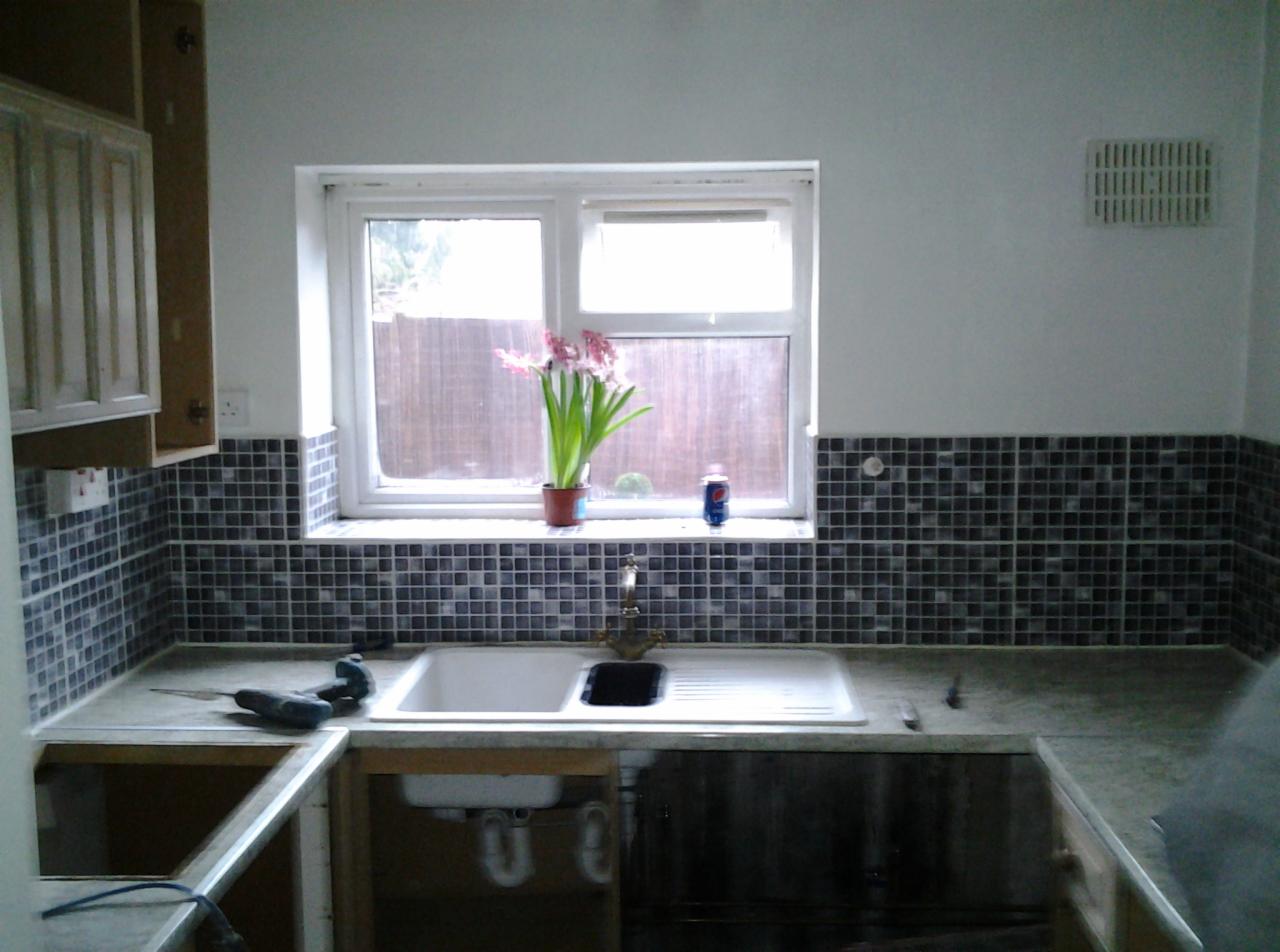 Częściowo wykończona kuchnia z mozaiką na ścianie, widoczny zlew, bateria, kwiaty w doniczce na parapecie i narzędzia na blacie.