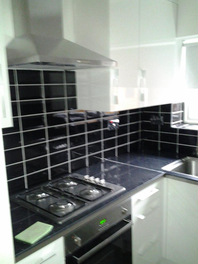 Czarno-biała kuchnia z błyszczącymi, czarnymi płytkami na ścianie nad blatem, czteropalnikową płytą gazową i srebrnym okapem.