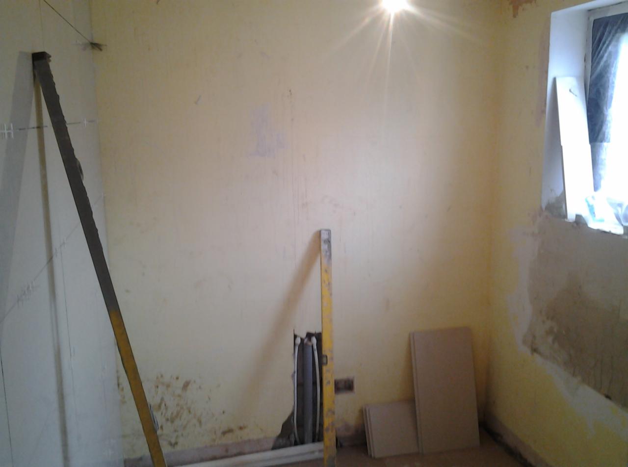 Wnętrze pokoju w trakcie przygotowań do remontu: widoczne piony wyznaczone sznurkiem, poziomica oparta o ścianę, odsłonięte rury instalacyjne i płyty na podłodze.