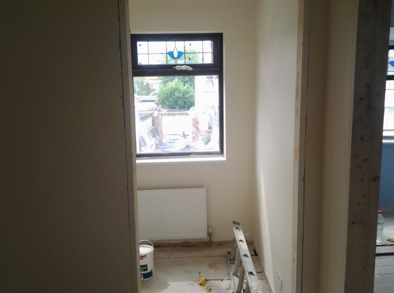 Wnętrze pokoju w trakcie remontu: świeżo pomalowane ściany na jasny kolor, okno z witrażem, grzejnik, drabina malarska i wiadro z farbą na podłodze.