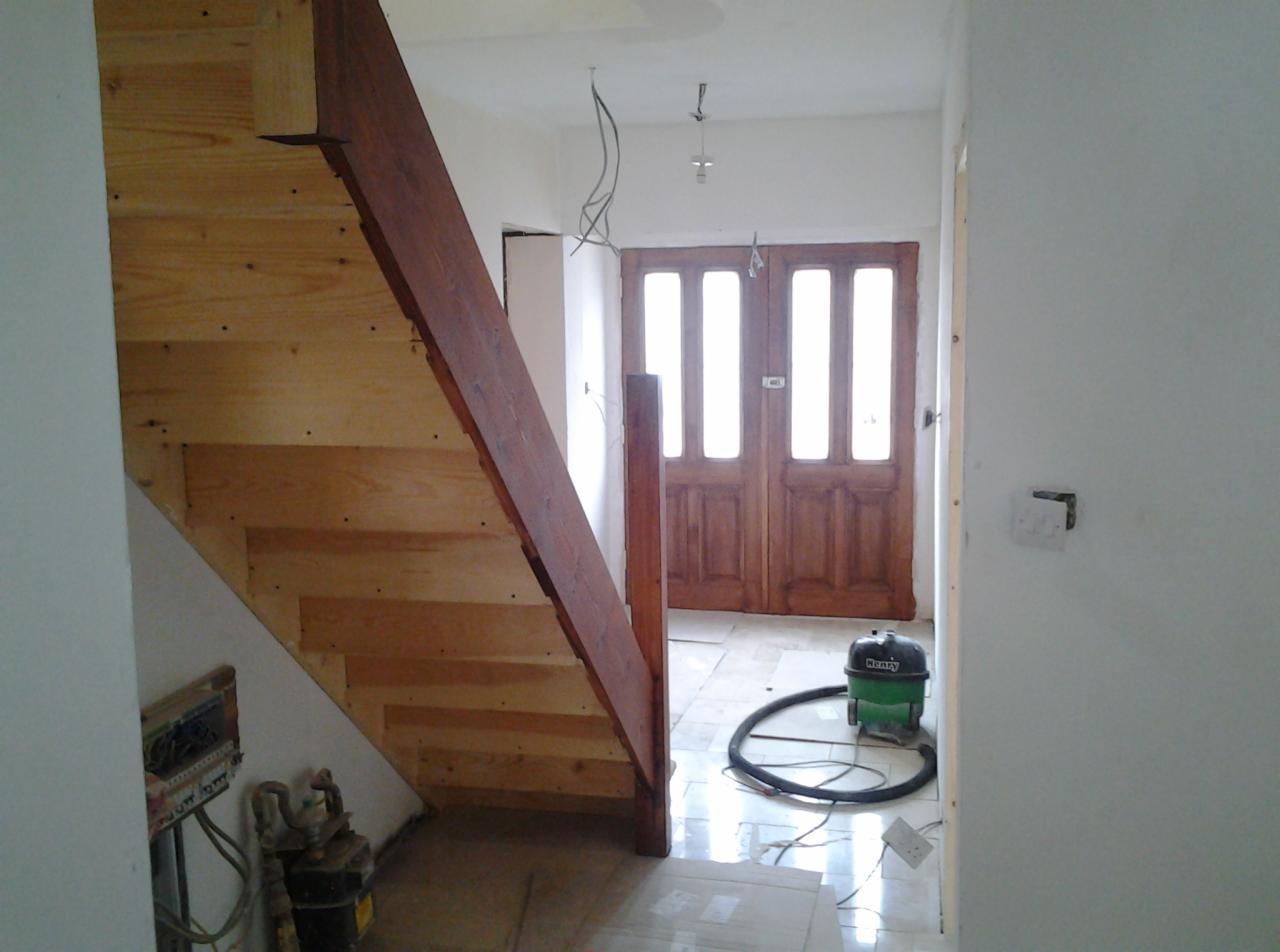 Surowe drewniane schody z ciemnobrązową poręczą w trakcie montażu, widoczny odkurzacz i przewody elektryczne, wnętrze w trakcie wykańczania.