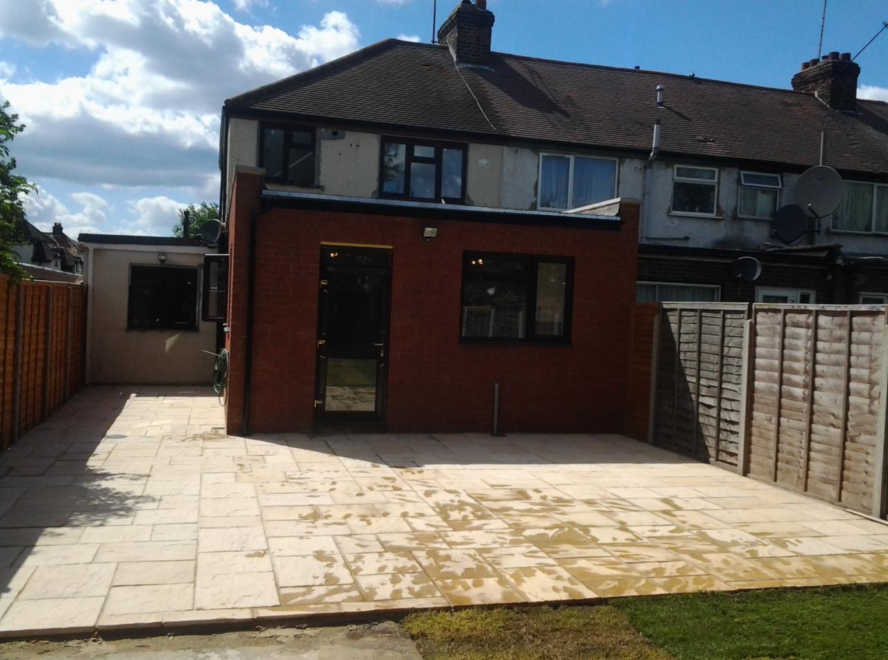 Nowo ułożona, jasna kostka brukowa na podwórku z ceglaną dobudówką i drewnianym ogrodzeniem, częściowo jeszcze mokra po myciu.